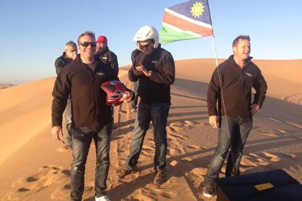 A group in branded jackets on the dunes at sunrise, a Namibian flag planted in the sand beside them
